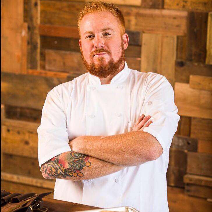 A man with beard and tattoos standing in front of wooden wall.