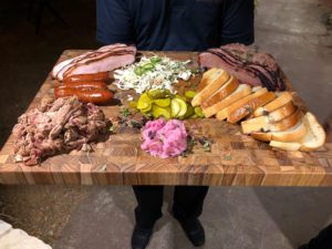 A person holding a wooden cutting board with meat and vegetables on it.
