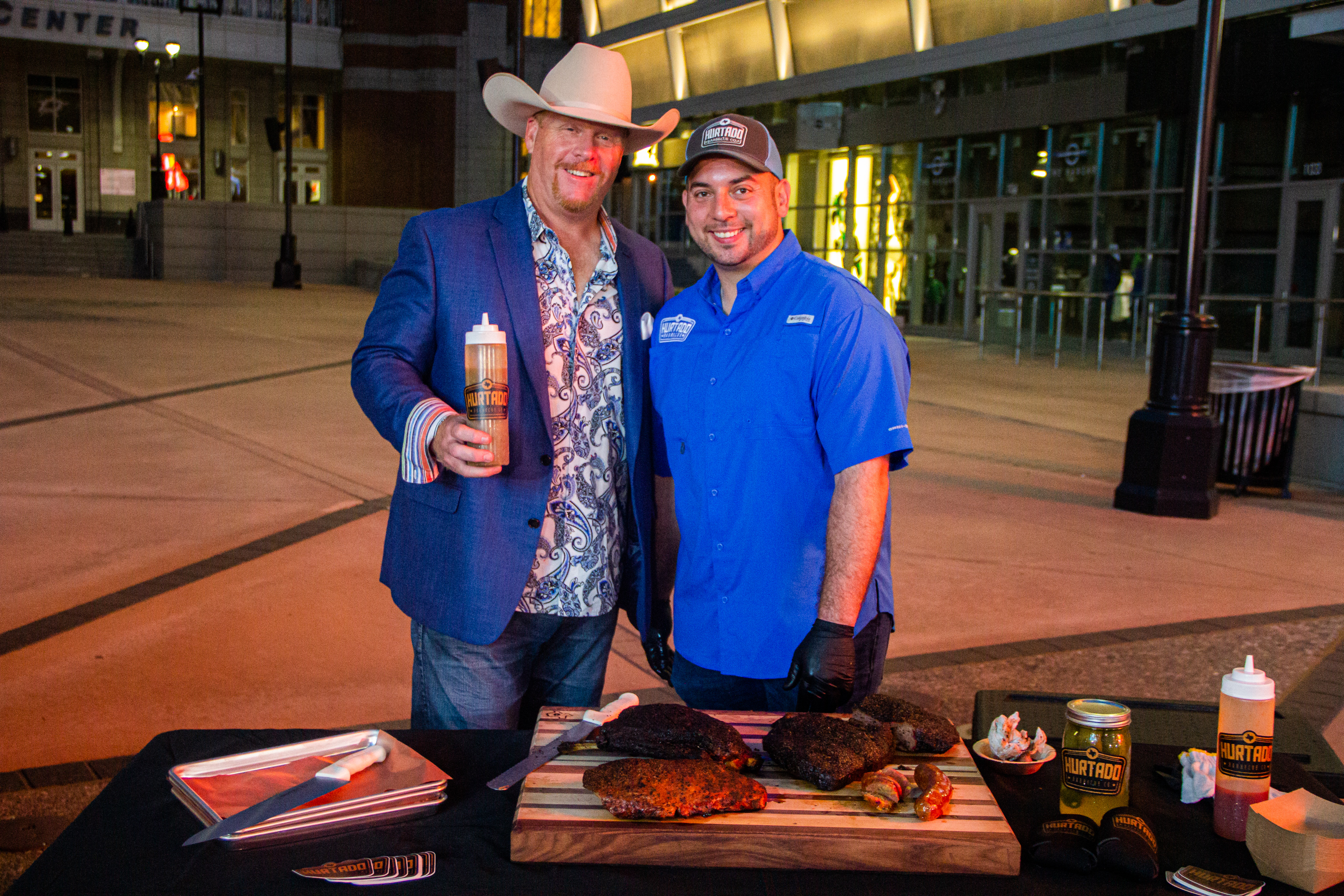 Two men standing next to a grill with meat on it.