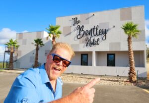 A man pointing to the camera in front of a hotel.