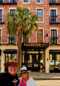 A man in a cowboy hat and sunglasses standing under a palm tree.